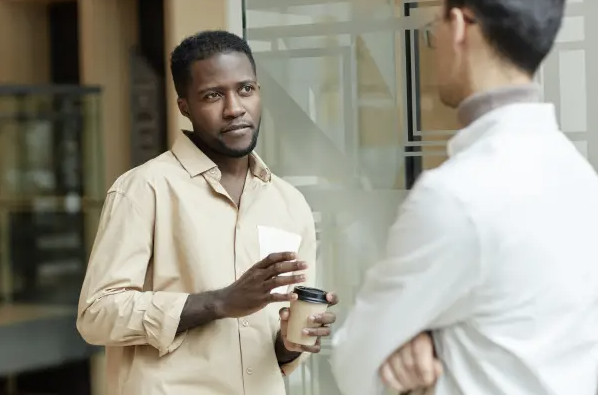 Two men stand indoors talking; one holds a coffee cup and wears a beige shirt.